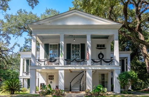 Large house painted white with dark shutters surrounded by green vegetation and large trees