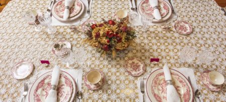 Dining table with delicate knitted tablecloth, Victorian red and white China plates, folded napkins, and red and yellow flower arrangement