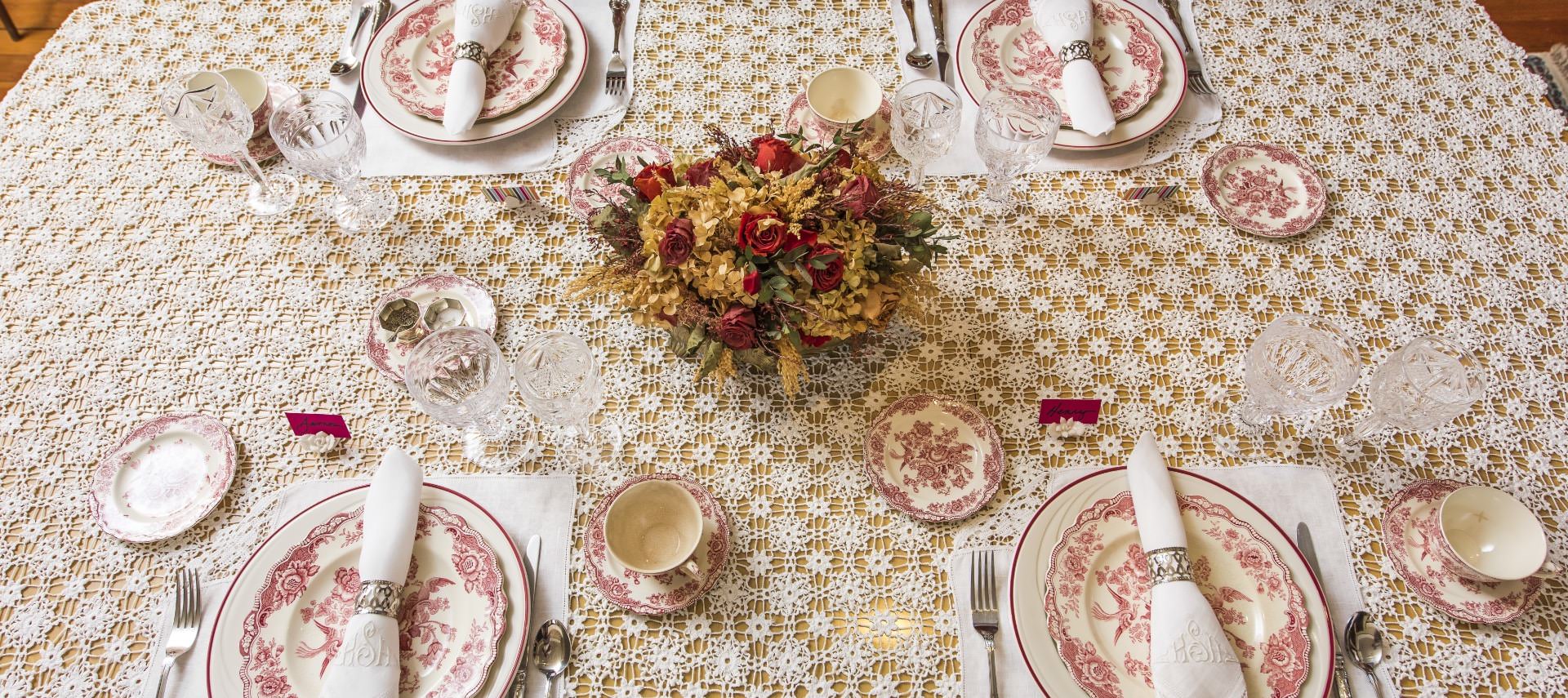 Dining table with delicate knitted tablecloth, Victorian red and white China plates, folded napkins, and red and yellow flower arrangement