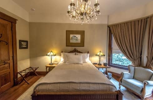 Bedroom with dark wooden bed, white bedding, wooden nightstands with antique lamps, Victorian chair, and chandelier