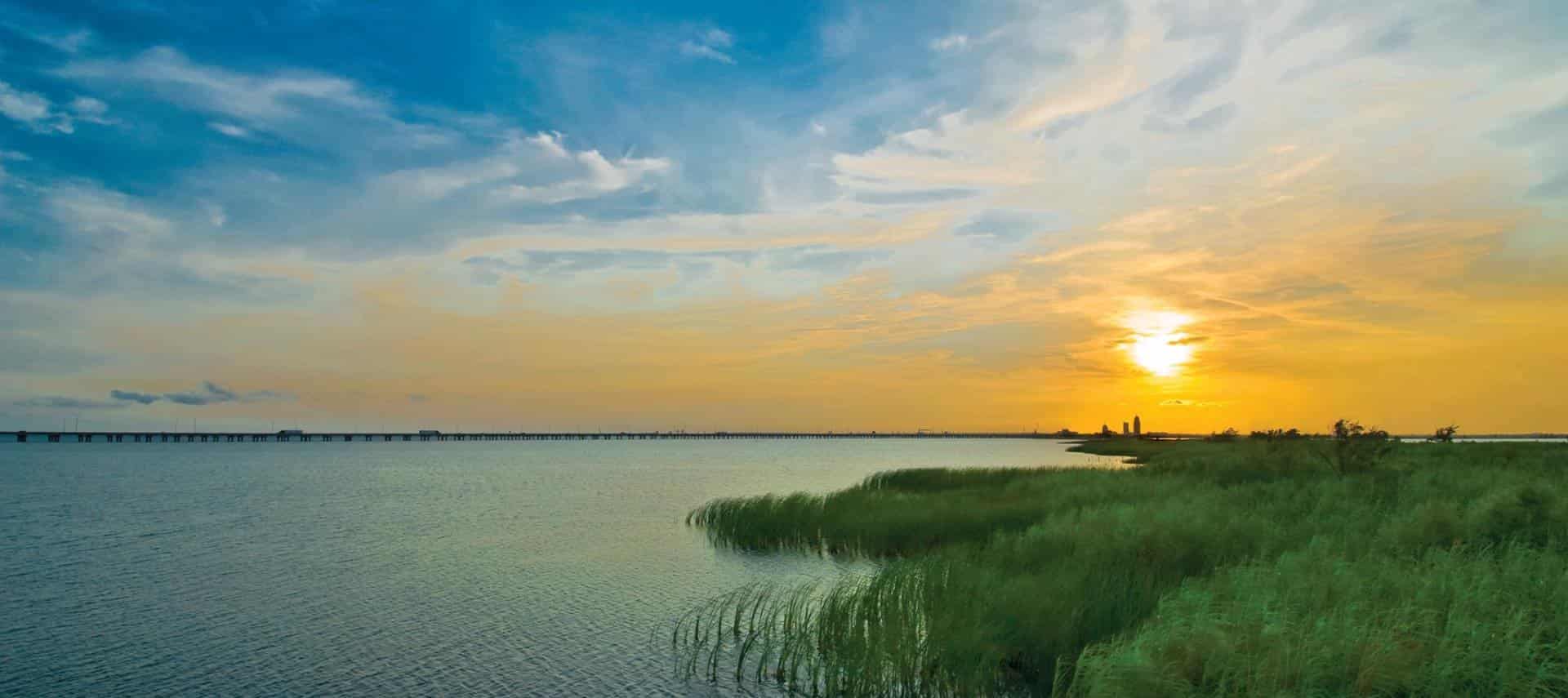 Sky with clouds at sunset with blue and yellow hues, long highway bridge over the water next to tall grass