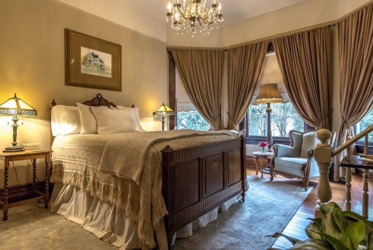 Bedroom with dark wooden bed, white bedding, wooden nightstands with antique lamps, Victorian chair, and chandelier