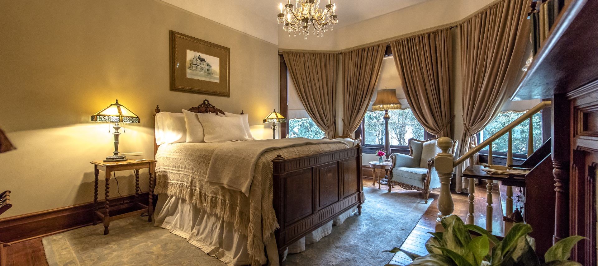 Bedroom with dark wooden bed, white bedding, wooden nightstands with antique lamps, Victorian chair, and chandelier