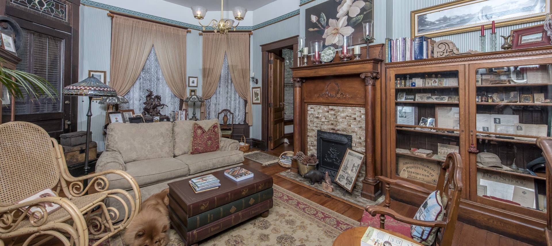 Living room with cream upholstered loveseat, wicker rocking chair, antique lamp, built in wooden bookcases, light brown brick fireplace with antique wooden mantel, and fluffy brown dog