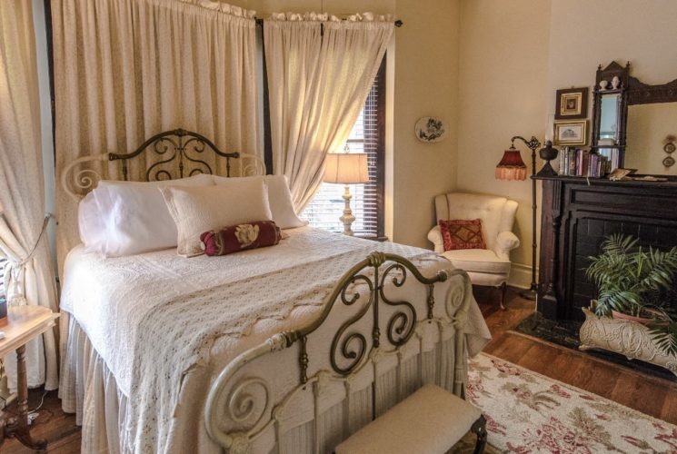Bedroom with old iron bedframe with white bedding, white upholstered chair in corner, and large black-bricked fireplace with dark wood mantel and mirror on top