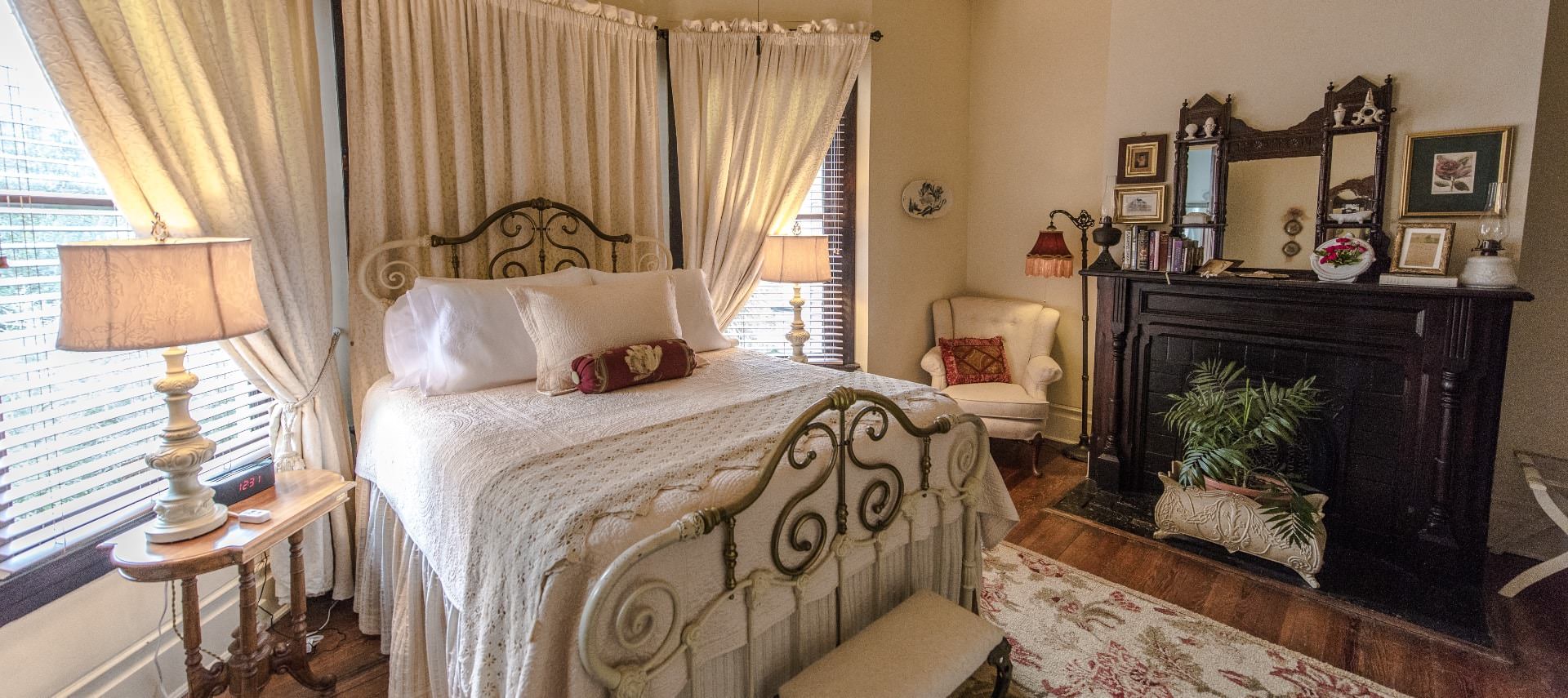 Bedroom with old iron bedframe with white bedding, white upholstered chair in corner, and large black-bricked fireplace with dark wood mantel and mirror on top